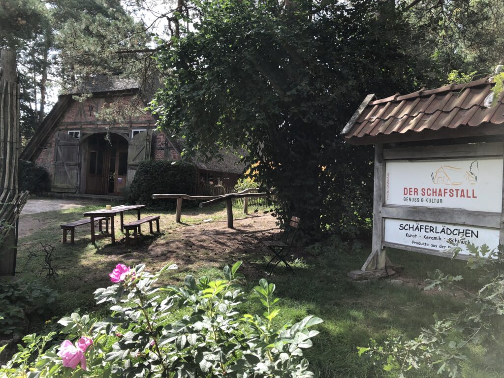 Ein Schild und im Hintergrund das Restaurant. Auf dem Schild ist der Name des Restaurants "Der Schafstall" zu lesen.